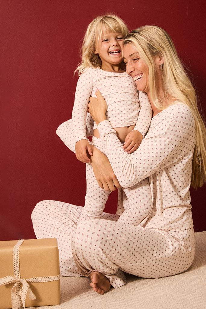 A smiling blonde woman and young child wearing matching cream pajamas with small red stars, sitting together against a dark red wall. A wrapped gift box is visible in the lower left corner.