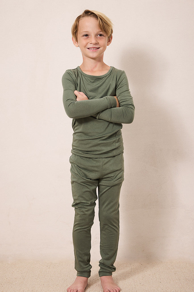 A young boy stands barefoot with arms crossed, wearing an olive green long-sleeve shirt and matching green pants against a plain beige studio background.