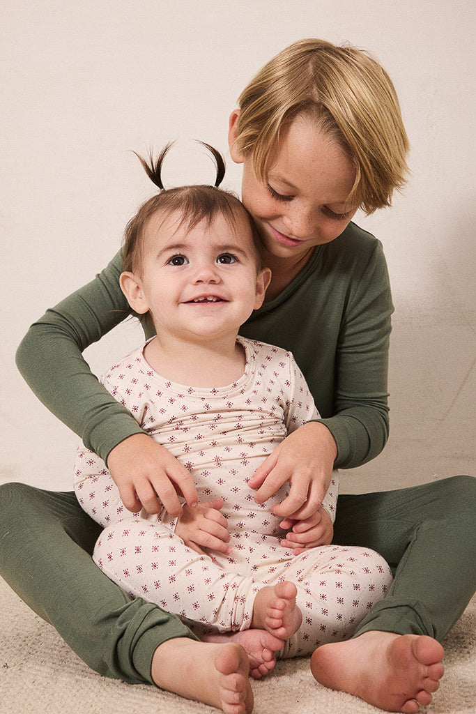 Two children sitting together on a light beige floor against a neutral beige wall. The older child has blonde hair and wears an olive green long-sleeve top and matching green pants, while the younger child wears a cream pajama top and matching pajama pants with a small red pattern.