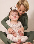 Two children sitting together on a light beige floor against a neutral beige wall. The older child has blonde hair and wears an olive green long-sleeve top and matching green pants, while the younger child wears a cream pajama top and matching pajama pants with a small red pattern.