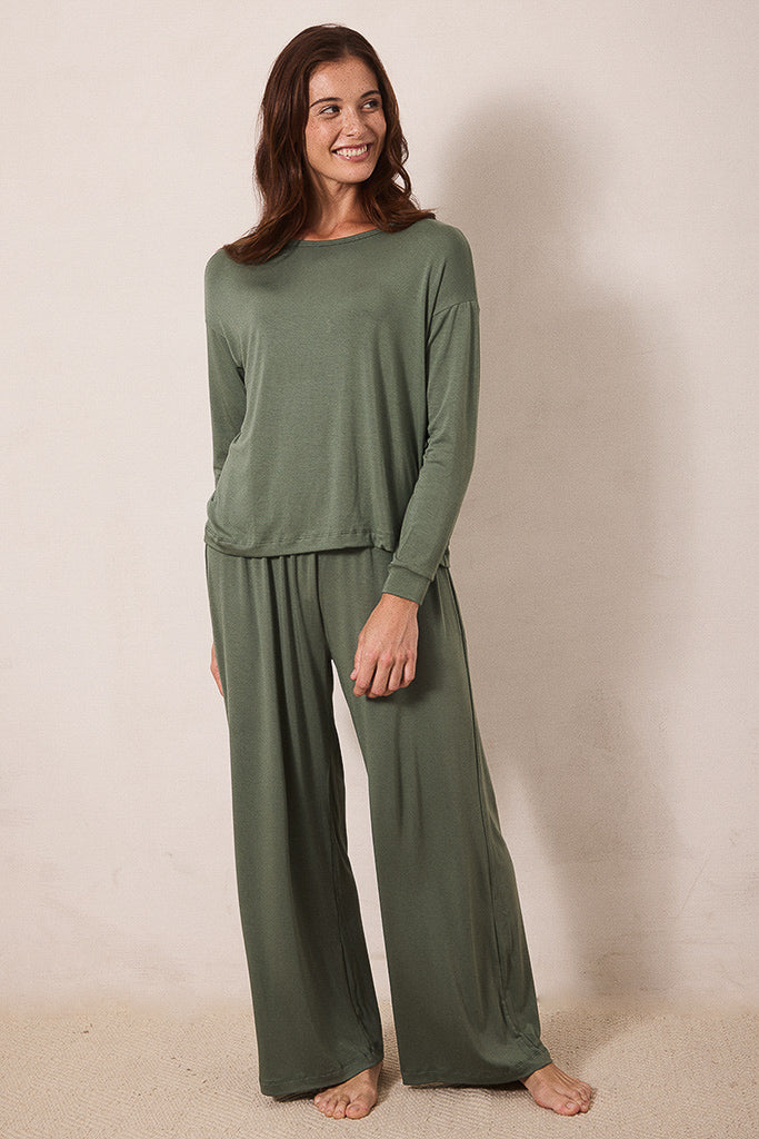 A woman modeling a matching two-piece lounge set in olive green, featuring a long-sleeve top and wide-leg pants, standing against a plain beige wall.