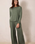 A woman modeling a matching two-piece lounge set in olive green, featuring a long-sleeve top and wide-leg pants, standing against a plain beige wall.