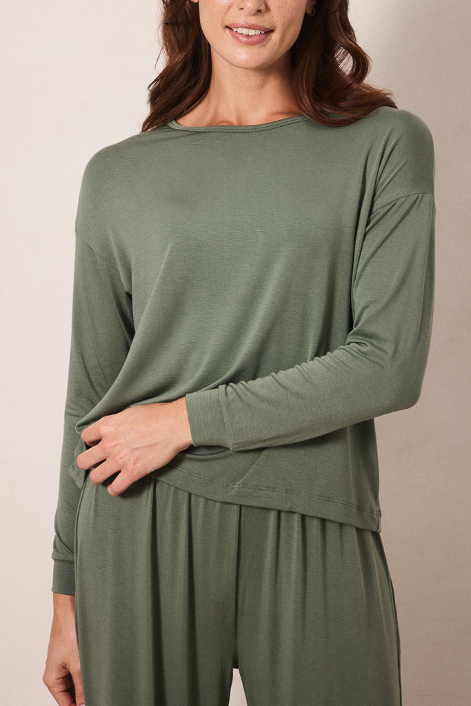 A woman wearing a sage green long-sleeve top and matching sage green pants, shown from the nose down against a plain beige studio background.