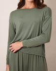 A woman wearing a sage green long-sleeve top and matching sage green pants, shown from the nose down against a plain beige studio background.