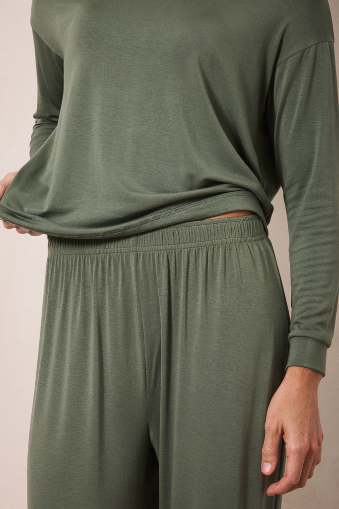 A close-up of a person wearing a matching two-piece lounge set in olive green, featuring a long-sleeve top and high-waisted pants with an elastic waistband.