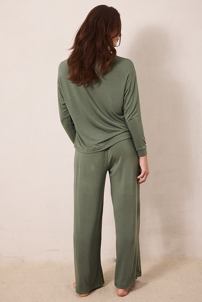 A woman viewed from behind wearing an olive green two-piece loungewear set consisting of a long-sleeve top and matching wide-leg pants, standing against a plain beige wall.