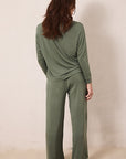 A woman viewed from behind wearing an olive green two-piece loungewear set consisting of a long-sleeve top and matching wide-leg pants, standing against a plain beige wall.