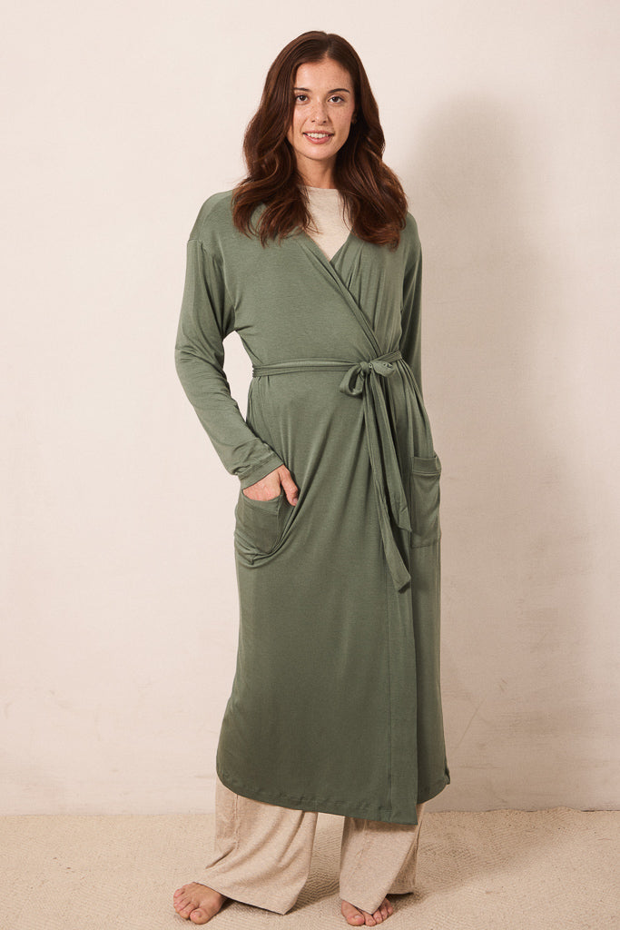 A woman wearing a long sage green wrap robe with a tie waist and front pockets, styled over a cream top and beige pants, standing against a plain beige wall.