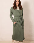 A woman wearing a long sage green wrap robe with a tie waist and front pockets, styled over a cream top and beige pants, standing against a plain beige wall.