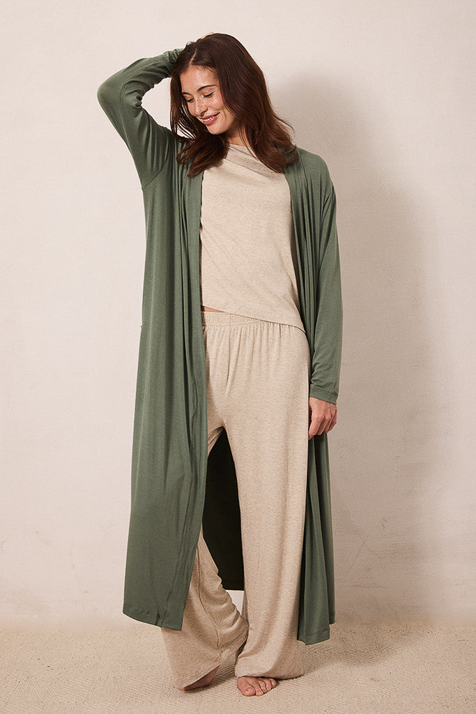 A woman modeling a long sage green open-front cardigan over a beige knit top and matching wide-leg pants, standing against a plain beige studio backdrop.
