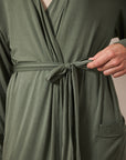 Close-up of a person wearing an olive green wrap-style robe or dress with a tie belt at the waist and a side pocket, against a plain light background.