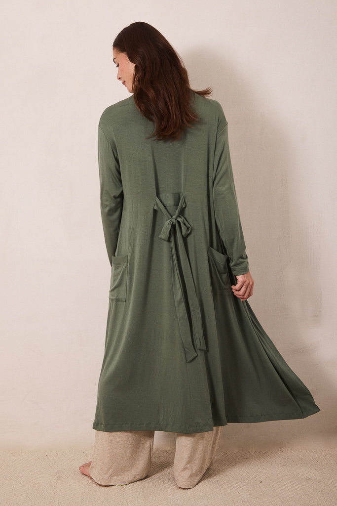 A woman shown from the back wearing a long olive green wrap-style cardigan or robe with a tie belt at the waist, paired with light beige pants, standing against a plain beige studio wall.