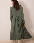 A woman shown from the back wearing a long olive green wrap-style cardigan or robe with a tie belt at the waist, paired with light beige pants, standing against a plain beige studio wall.