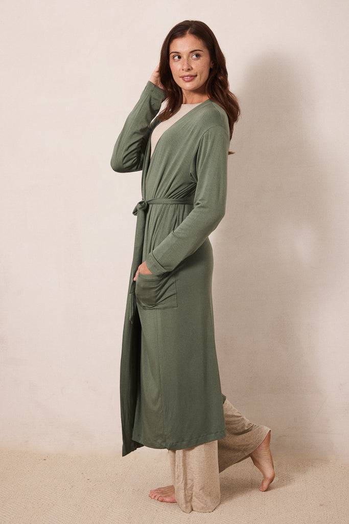 A woman modeling a long-sleeve sage green robe with a tie waist and front pockets, worn over a cream top and beige pants, standing against a plain beige studio wall.