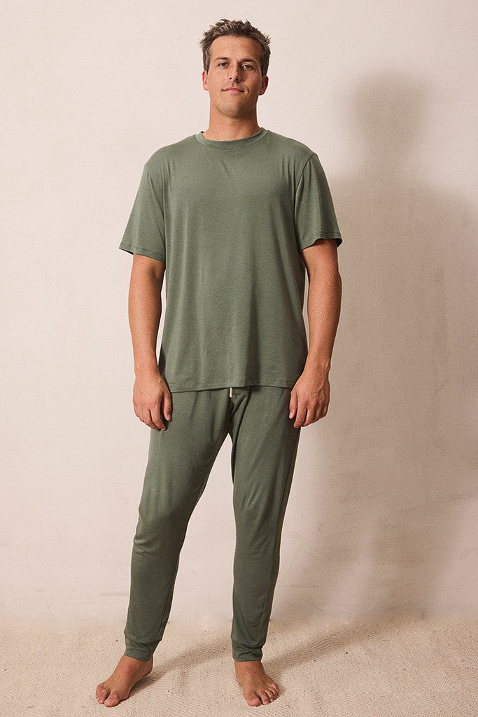 A man modeling a matching two-piece pajama set in olive green, consisting of a short-sleeve crewneck t-shirt and matching jogger pants, standing against a plain beige wall.