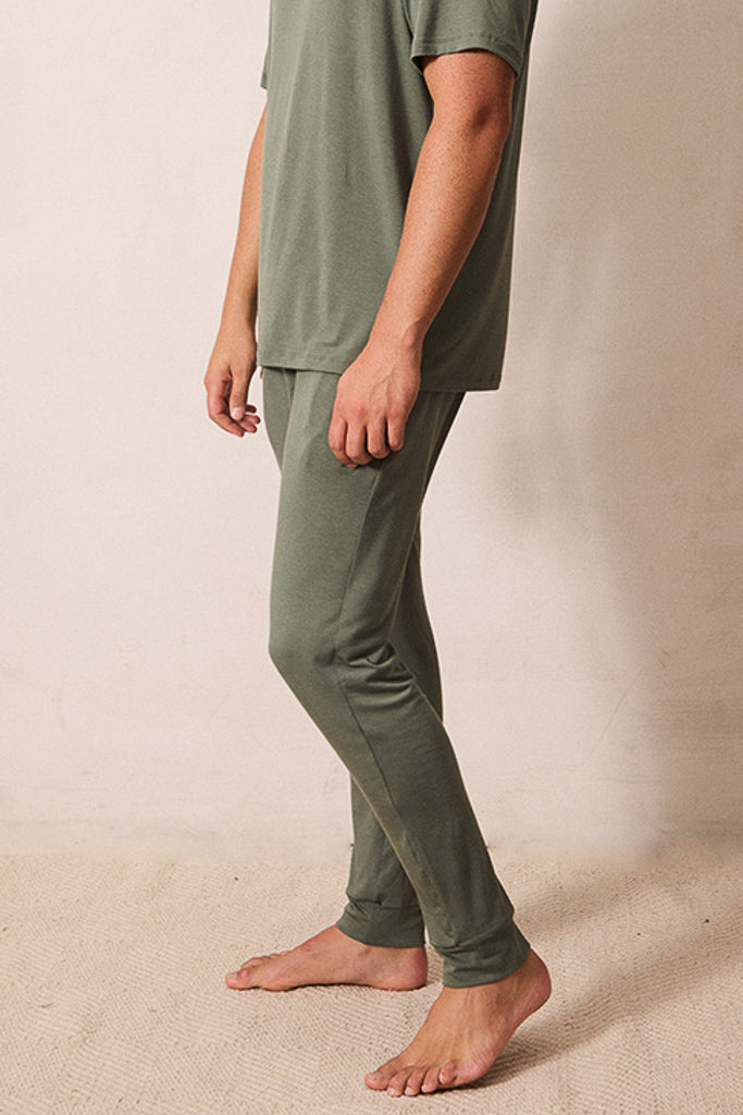 A person wearing a matching two-piece lounge set in olive green, featuring a short-sleeve crewneck t-shirt and slim-fit pants, standing barefoot against a neutral beige wall.