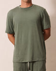 A man wearing a matching olive green short-sleeve t-shirt and sweatpants set, standing against a plain beige background.
