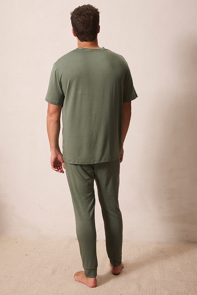 A man viewed from behind wearing an olive green short-sleeve t-shirt and matching olive green pajama pants, standing barefoot against a plain beige wall.
