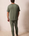 A man viewed from behind wearing an olive green short-sleeve t-shirt and matching olive green pajama pants, standing barefoot against a plain beige wall.