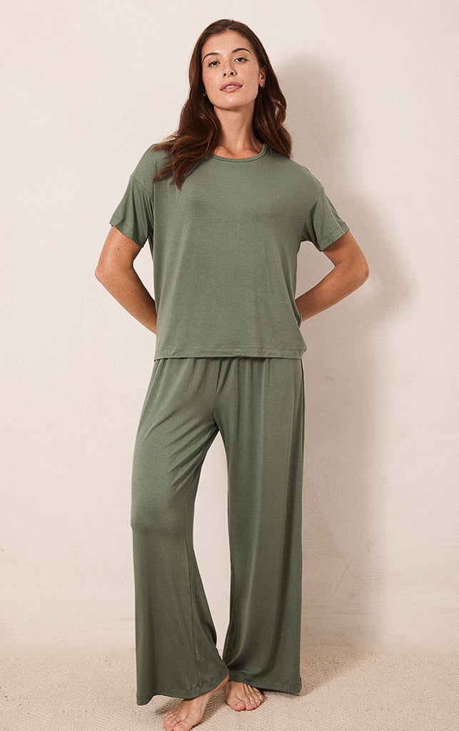 A woman modeling a matching two-piece lounge set in sage green, featuring a short-sleeve crewneck top and wide-leg pants, standing against a plain beige wall.