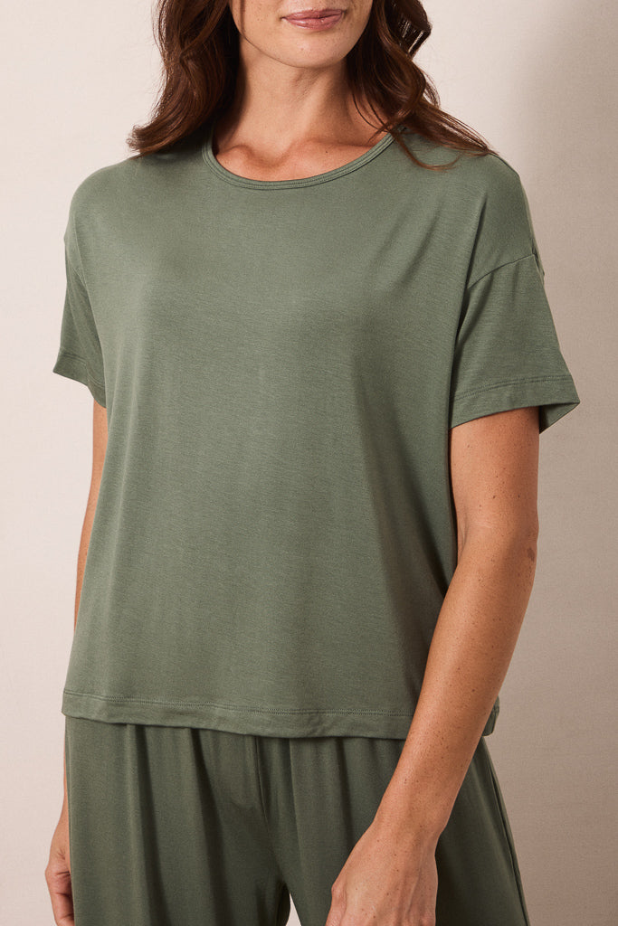 A woman wearing a loose, short-sleeve crewneck t-shirt in sage green, paired with matching sage green pants, photographed against a neutral beige background.