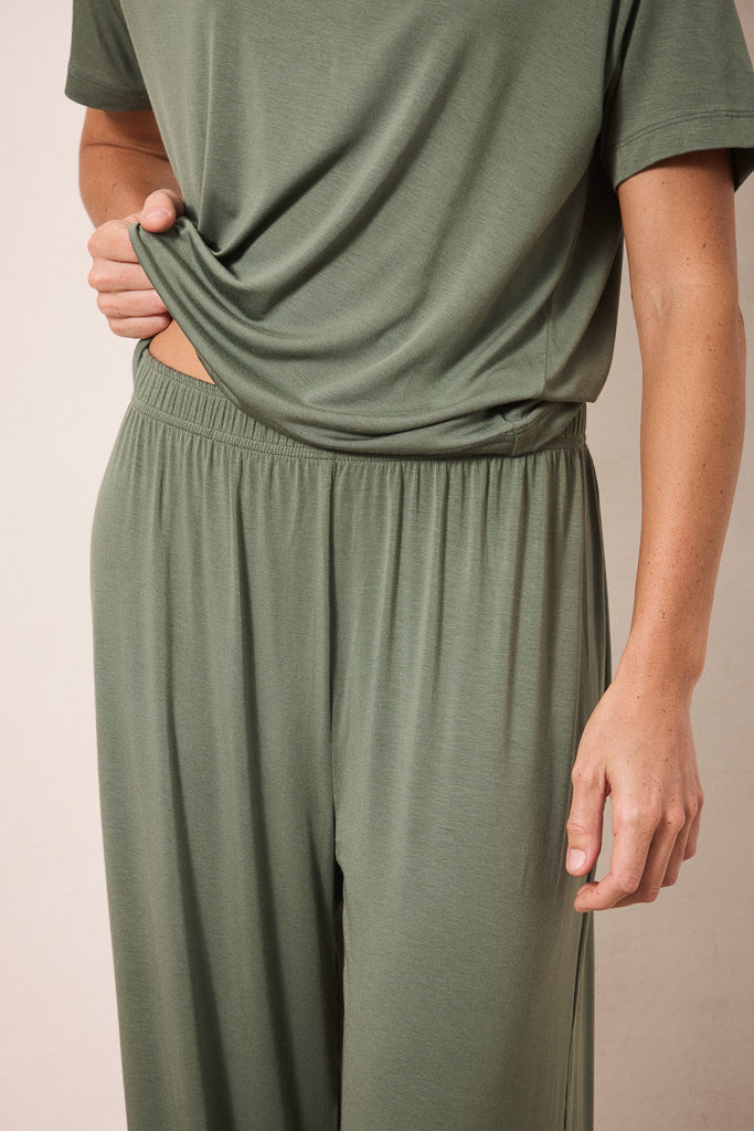 A close-up of a person wearing an olive green lounge set, featuring a short-sleeve top and matching wide-leg pants, with the hand lifting the hem of the shirt.