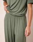 A close-up of a person wearing an olive green lounge set, featuring a short-sleeve top and matching wide-leg pants, with the hand lifting the hem of the shirt.