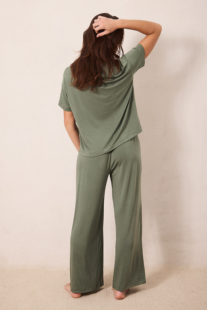 A woman wearing an olive green two-piece lounge set consisting of a short-sleeve top and wide-leg pants, shown from behind against a plain beige wall.