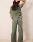 A woman wearing an olive green two-piece lounge set consisting of a short-sleeve top and wide-leg pants, shown from behind against a plain beige wall.