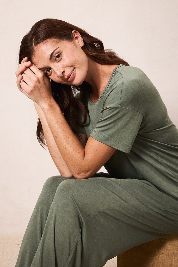 A woman wearing an olive green short-sleeve pajama set, sitting on a wooden stool against a neutral beige background.