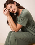 A woman wearing an olive green short-sleeve pajama set, sitting on a wooden stool against a neutral beige background.