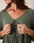 Close-up of a woman wearing an olive green V-neck dress with tie-front detailing and gathered sleeves, shown against a plain beige background.
