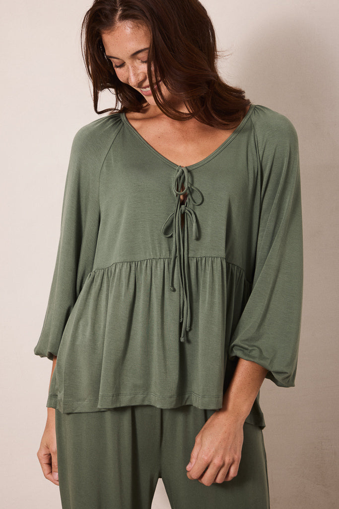 A woman wearing an olive green long-sleeve top with a V-neck, tie-front detail, and gathered waist, paired with matching green pants, against a plain beige studio background.