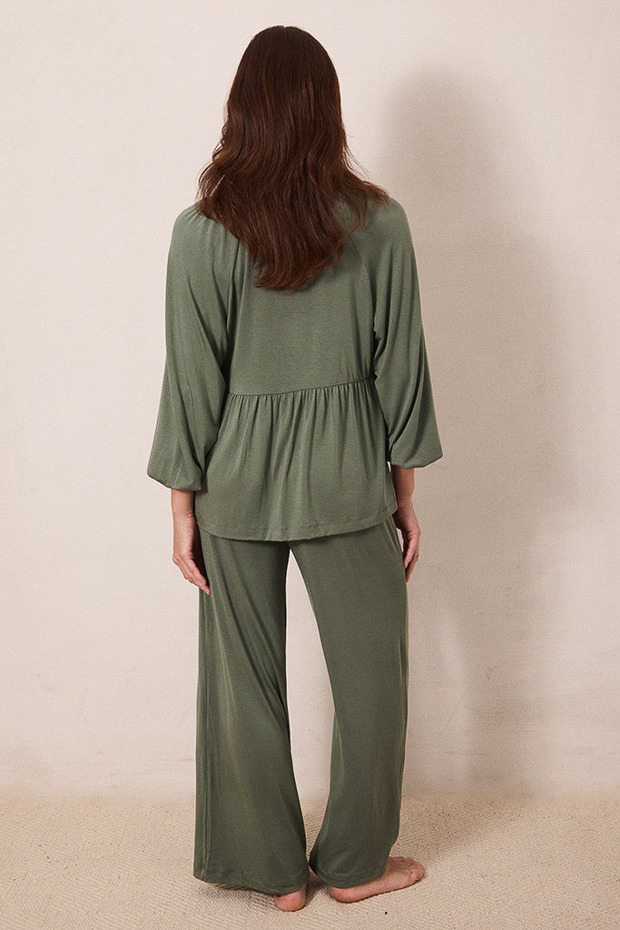 A woman viewed from behind wearing an olive green two-piece lounge set consisting of a long-sleeve peplum top and matching wide-leg pants, standing barefoot against a plain beige wall.
