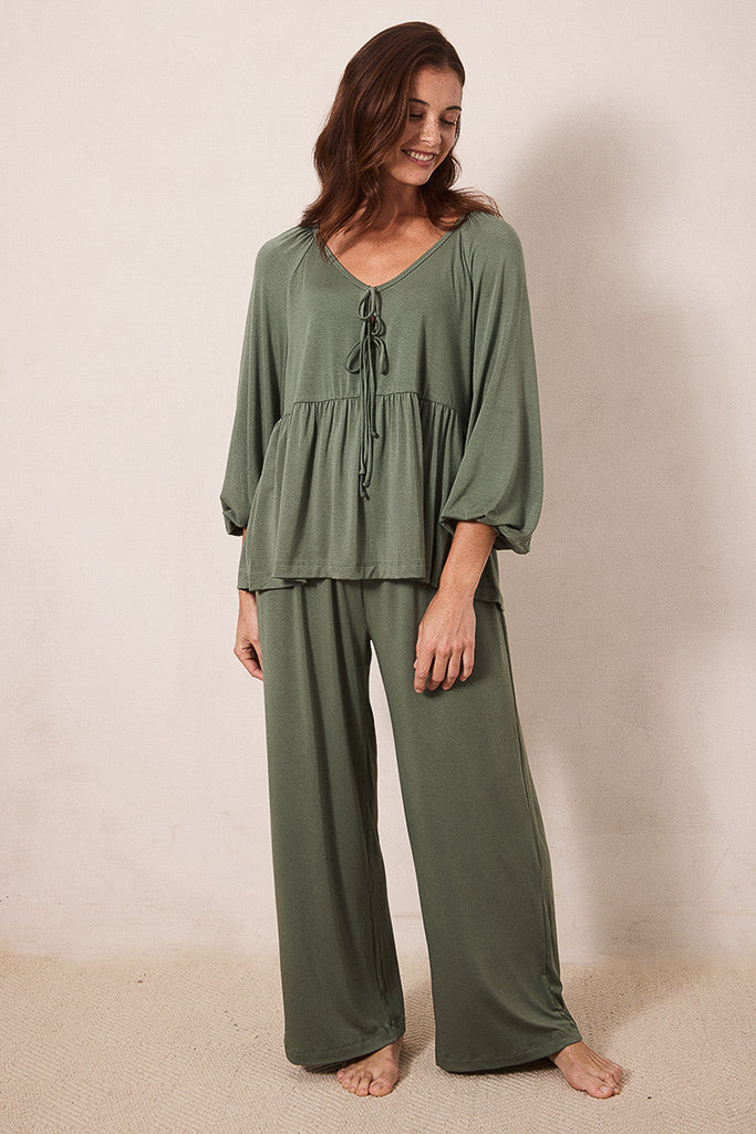 A woman modeling a matching two-piece lounge set in sage green, featuring a long-sleeve tie-front top and wide-leg pants, standing against a plain beige wall.