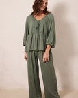 A woman modeling a matching two-piece lounge set in sage green, featuring a long-sleeve tie-front top and wide-leg pants, standing against a plain beige wall.