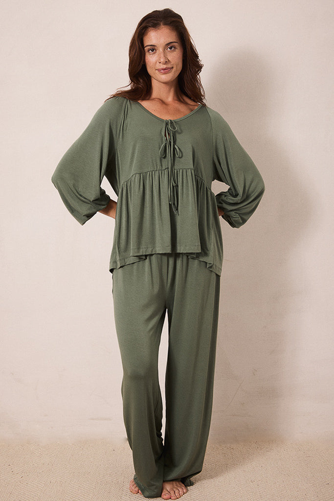 A woman modeling a matching two-piece lounge set in olive green, featuring a loose long-sleeve top with a tie neckline and matching wide-leg pants, set against a plain beige studio background.
