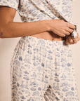 A close-up of a person wearing a matching short-sleeve top and pants pajama set with a blue toile de Jouy pattern on a cream background.