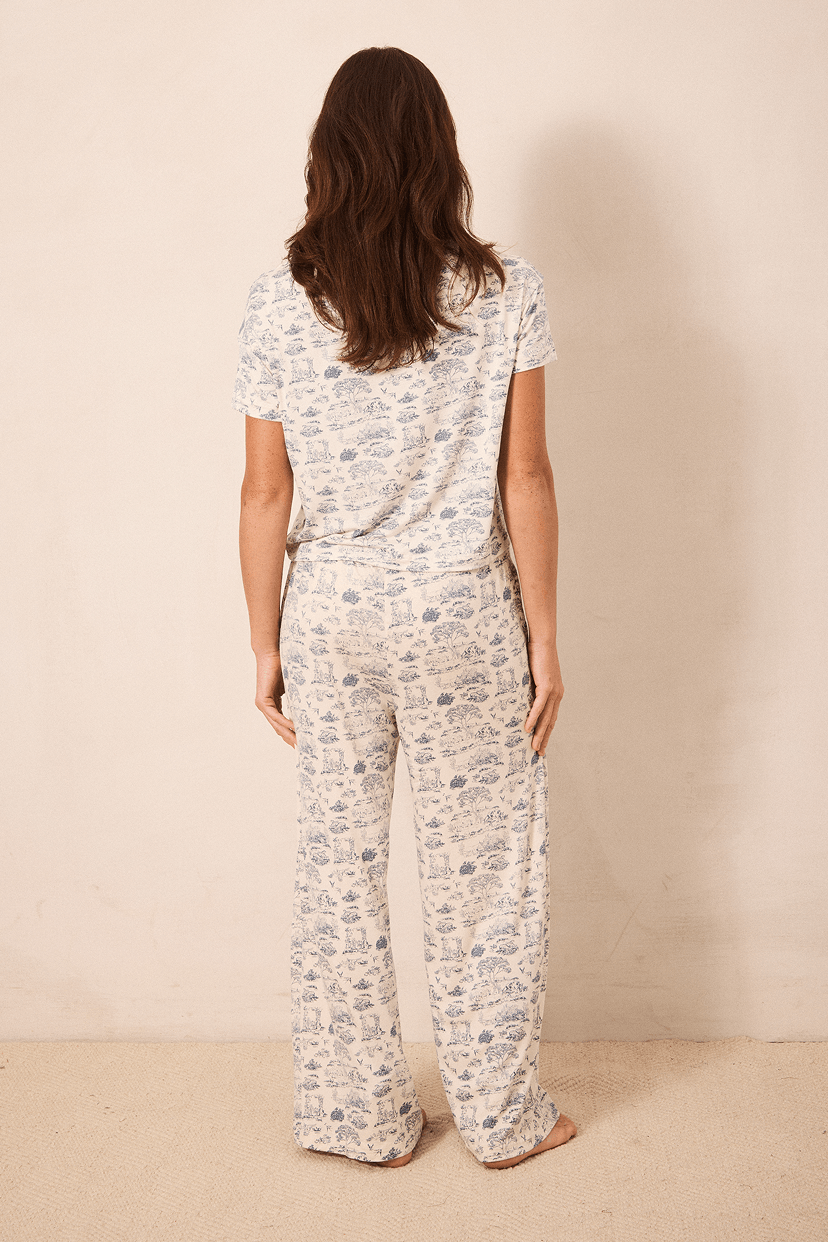 A woman viewed from behind wearing a matching short-sleeve top and wide-leg pants pajama set with a blue toile pattern on a cream background, standing against a plain beige wall.