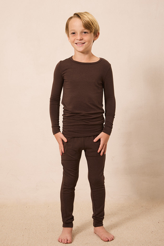 A young boy with blonde hair stands barefoot against a warm beige studio background, wearing a matching dark brown long-sleeve top and pants set.