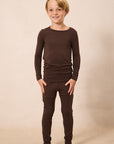 A young boy with blonde hair stands barefoot against a warm beige studio background, wearing a matching dark brown long-sleeve top and pants set.