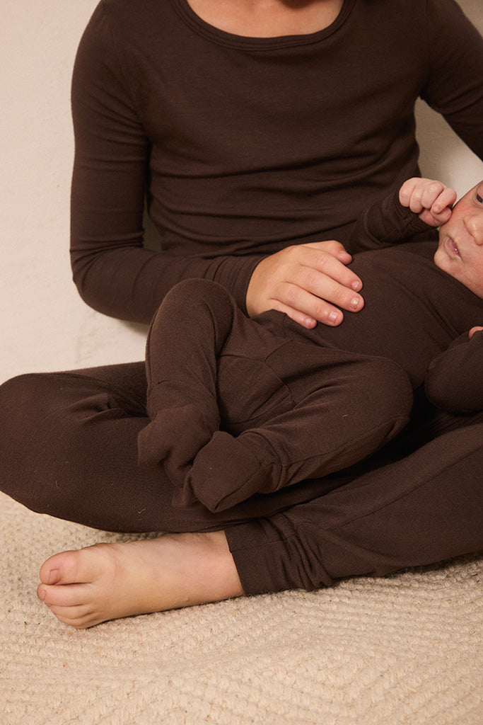 A big sibling and a baby wearing matching dark brown long-sleeve pajamas, sitting on a beige textured carpet.