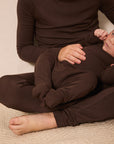 A big sibling and a baby wearing matching dark brown long-sleeve pajamas, sitting on a beige textured carpet.