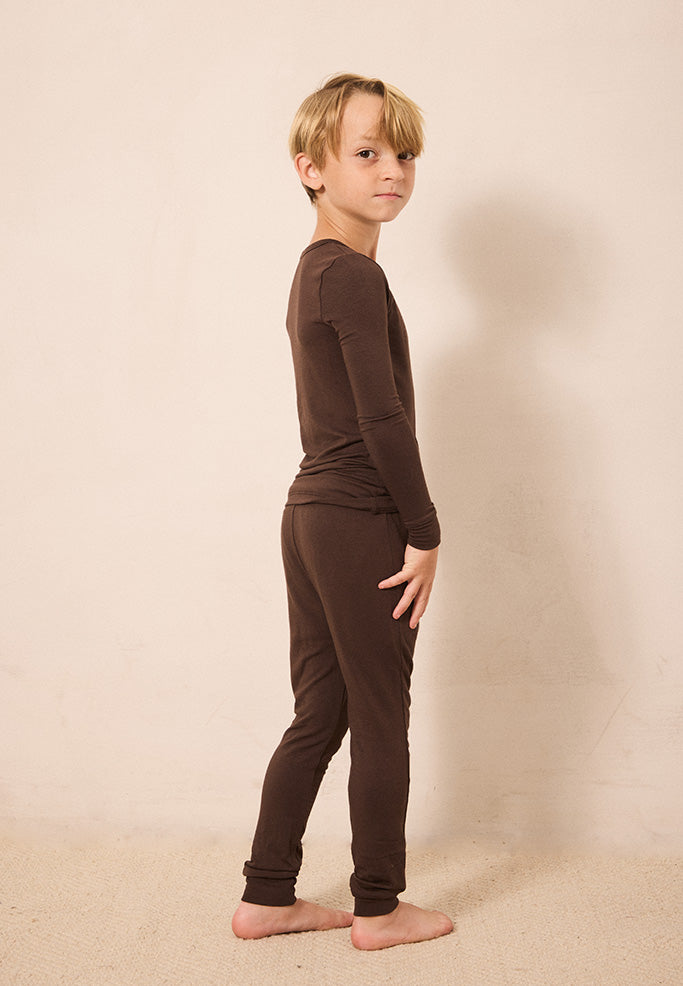 A young boy with blonde hair stands in profile wearing a matching dark brown long-sleeve top and jogger pants set, barefoot against a plain beige studio backdrop.