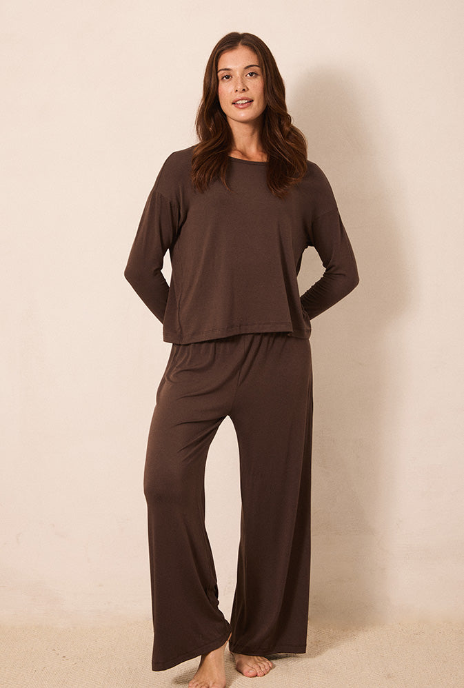 A woman modeling a dark brown two-piece loungewear set consisting of a long-sleeve crewneck top and matching wide-leg pants, standing against a plain beige studio backdrop.