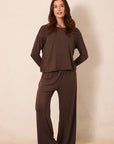 A woman modeling a dark brown two-piece loungewear set consisting of a long-sleeve crewneck top and matching wide-leg pants, standing against a plain beige studio backdrop.
