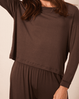 A woman wearing a dark brown long-sleeve top and matching brown pants against a plain beige studio background.