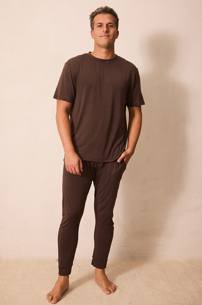 A man wearing a matching dark brown short-sleeve t-shirt and jogger pants set, standing barefoot against a plain beige wall.