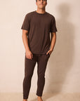 A man wearing a matching dark brown short-sleeve t-shirt and jogger pants set, standing barefoot against a plain beige wall.