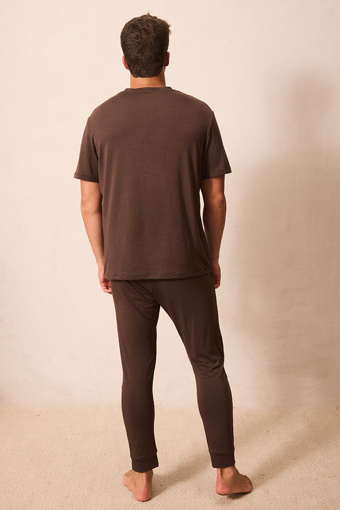 A man viewed from behind wearing a matching dark brown short-sleeve t-shirt and long pants pajama set, standing barefoot against a plain beige studio background.